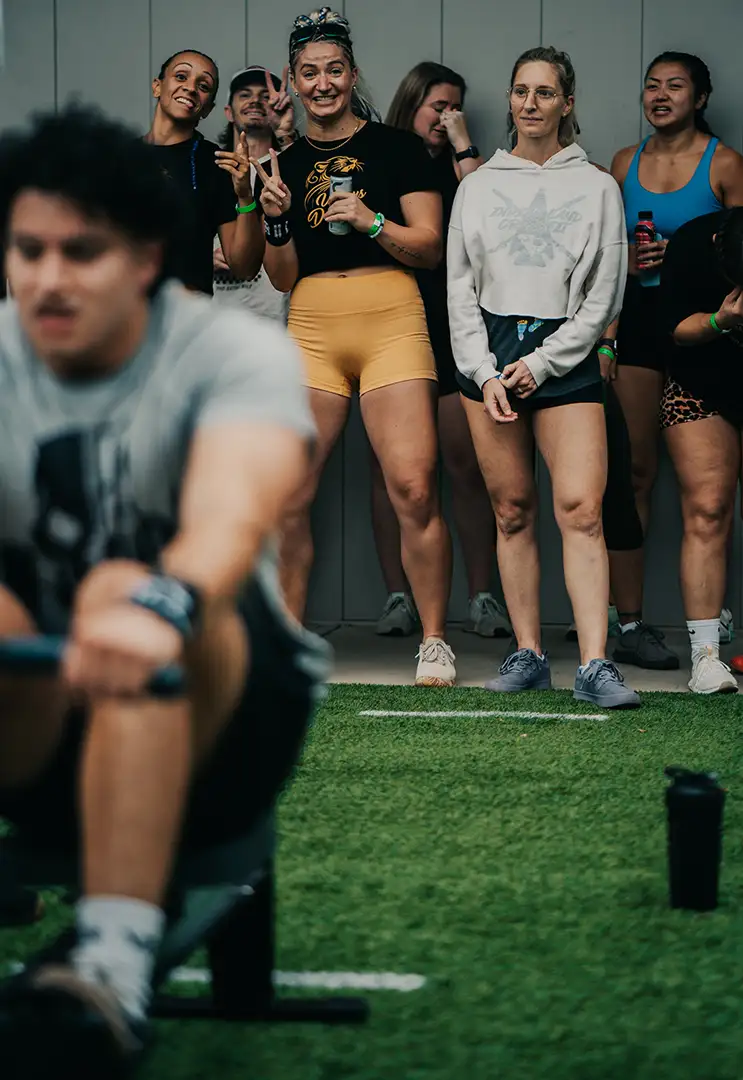 A young man competes on rowing machine while a group of young women watch in the background.